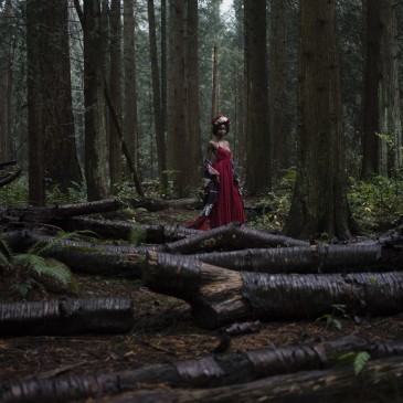 Natural light portrait of a young woman in a red dress and flower crown standing in the forest surrounded by fallen logs in Pacific Spirit National Park in Vancouver BC