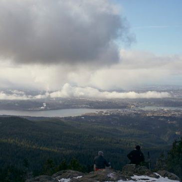 Image of 2 people sitting on rocks in the foreground looking across the Vancouver skyline from the top of Dog Mountain on a partly cloudy day