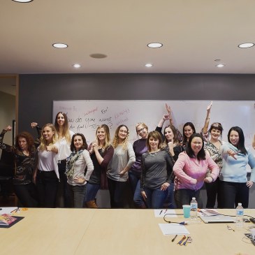 Image of a group of women in power poses in front of a whiteboard with empowering phrases during a self care workshop in Vancouver